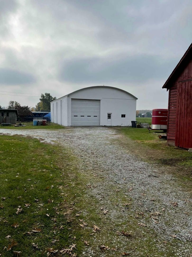 A white building with a garage door is sitting in the middle of a grassy field next to a red barn.