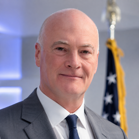 Portrait of a bald man in a gray suit and blue tie, standing before a blurred U.S. flag
