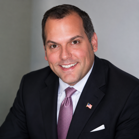 Smiling man in a dark suit, white shirt, and pink tie with an American flag pin on a gray background