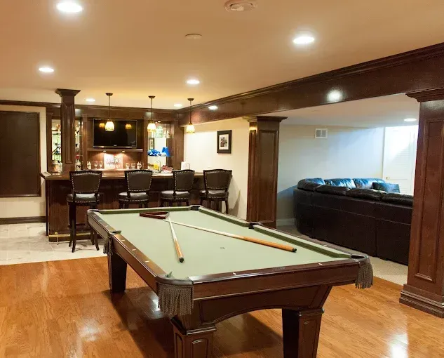 Game room with pool table, bar, seating area. Wooden details, warm lighting.