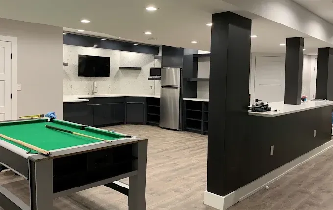 Basement with pool table, kitchen area, and dark columns. Grey walls, light wood floor.