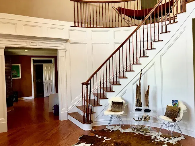 Elegant foyer with wooden staircase, white walls, and wood floor. Two chairs and a cowhide rug.