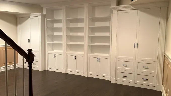 Built-in white bookshelves and cabinets in a basement.  Dark wood floor, stairs and light walls.