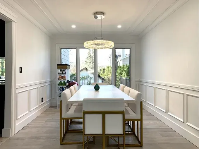 Formal dining room with a white table, chairs, and ornate wall trim, overlooking a window.