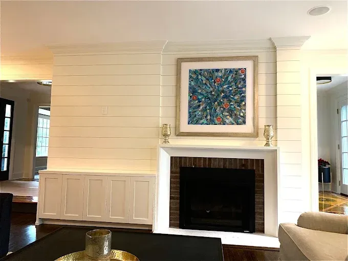 Fireplace with painting and built-in storage. Off-white paneling, black fireplace, gold accents, bright artwork.