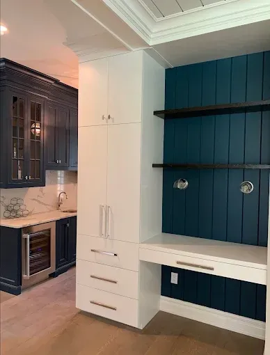 Built-in white storage unit and desk with navy blue accent wall, cabinetry in kitchen.