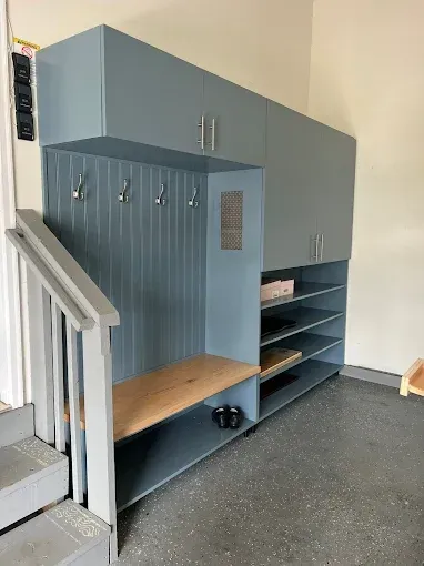 Blue built-in mudroom with cabinets, shelves, coat hooks, bench, and stairs.