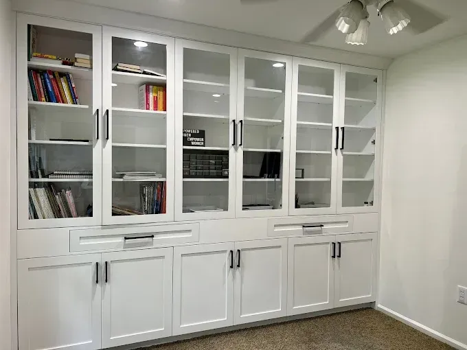 White built-in bookcase with glass doors, lower cabinets, and black hardware against a beige wall.