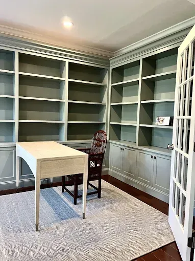 Home office with built-in green bookshelves, desk, patterned chair, and rug; French doors open to outside.