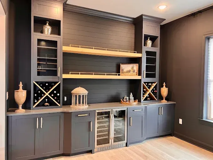 Built-in dark gray bar with wine racks, shelves, and a refrigerator in a room with light wood floors.