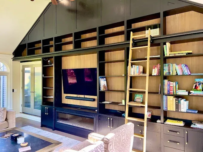 Built-in black bookcase with a ladder, TV, and fireplace. Light wood interior and books.