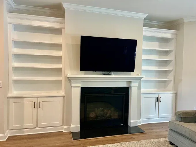 Built-in white bookshelves flanking a fireplace with a mounted TV. Beige walls, hardwood floor.