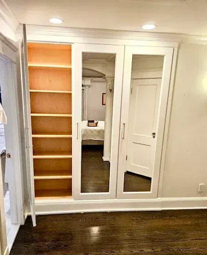 Built-in white closet with mirrored doors and open bookshelf on dark wood floor.