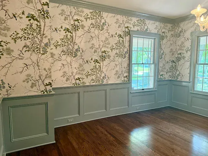Room with botanical wallpaper, pale green wainscoting, and hardwood floors. Two windows.