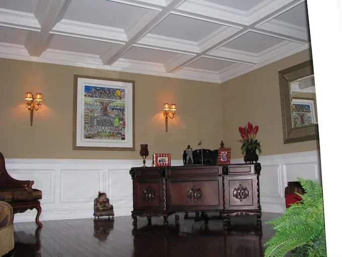 Office with dark wood desk, beige walls, wainscoting, coffered ceiling, artwork, sconces, and dark wood floors.