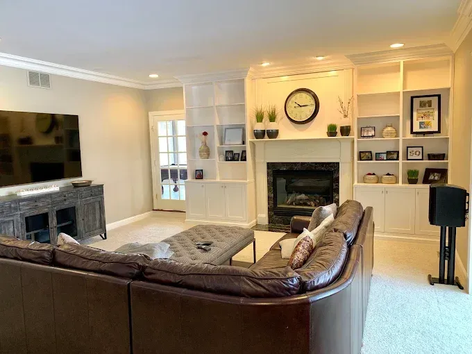 Living room with brown leather sectional, fireplace, built-in shelving, large TV, and light-colored walls.