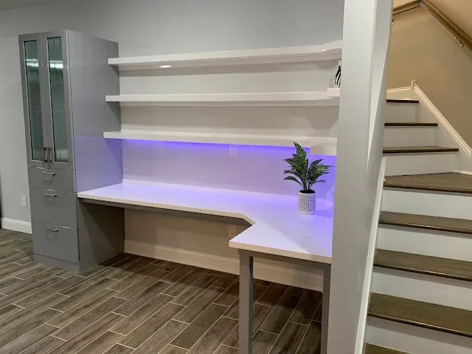 L-shaped white desk with shelves and purple under-lighting, next to a gray cabinet and a staircase.