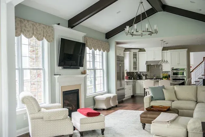 Living room with fireplace, TV, and open kitchen; light blue walls, wood beams, white furniture.