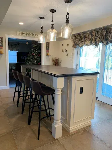 Kitchen island with 4 bar stools, 3 pendant lights above. White cabinetry, dark countertop, & glass door.