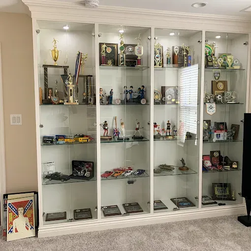 Four white display cabinets filled with trophies and collectibles, in a room with beige walls and carpet.