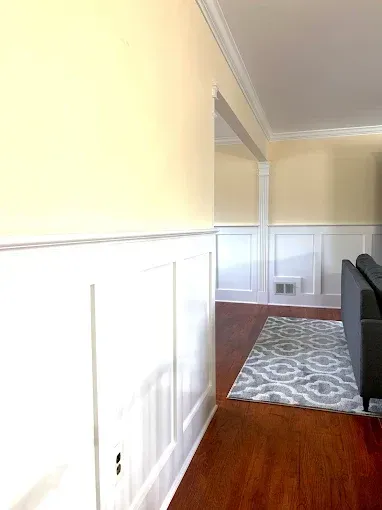 White wainscoting and trim adorn a room with hardwood floors, a gray rug, and a yellow wall.