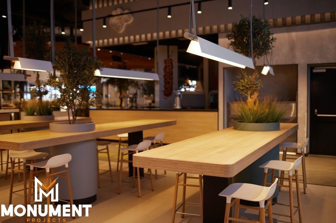 Modern indoor cafe with two long, light-wood communal tables, white bar stools, greenery, and hanging light fixtures.