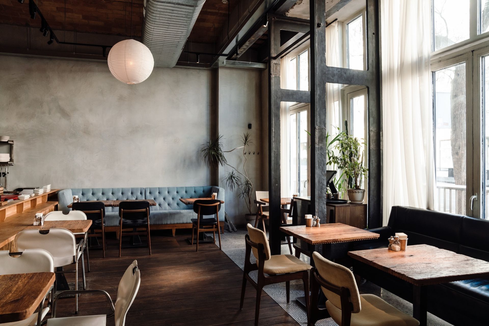 Modern Restaurant Interior Featuring Wooden Tables, Light-Colored Chairs, a Blue Tufted Booth, and Large Window Curtains.