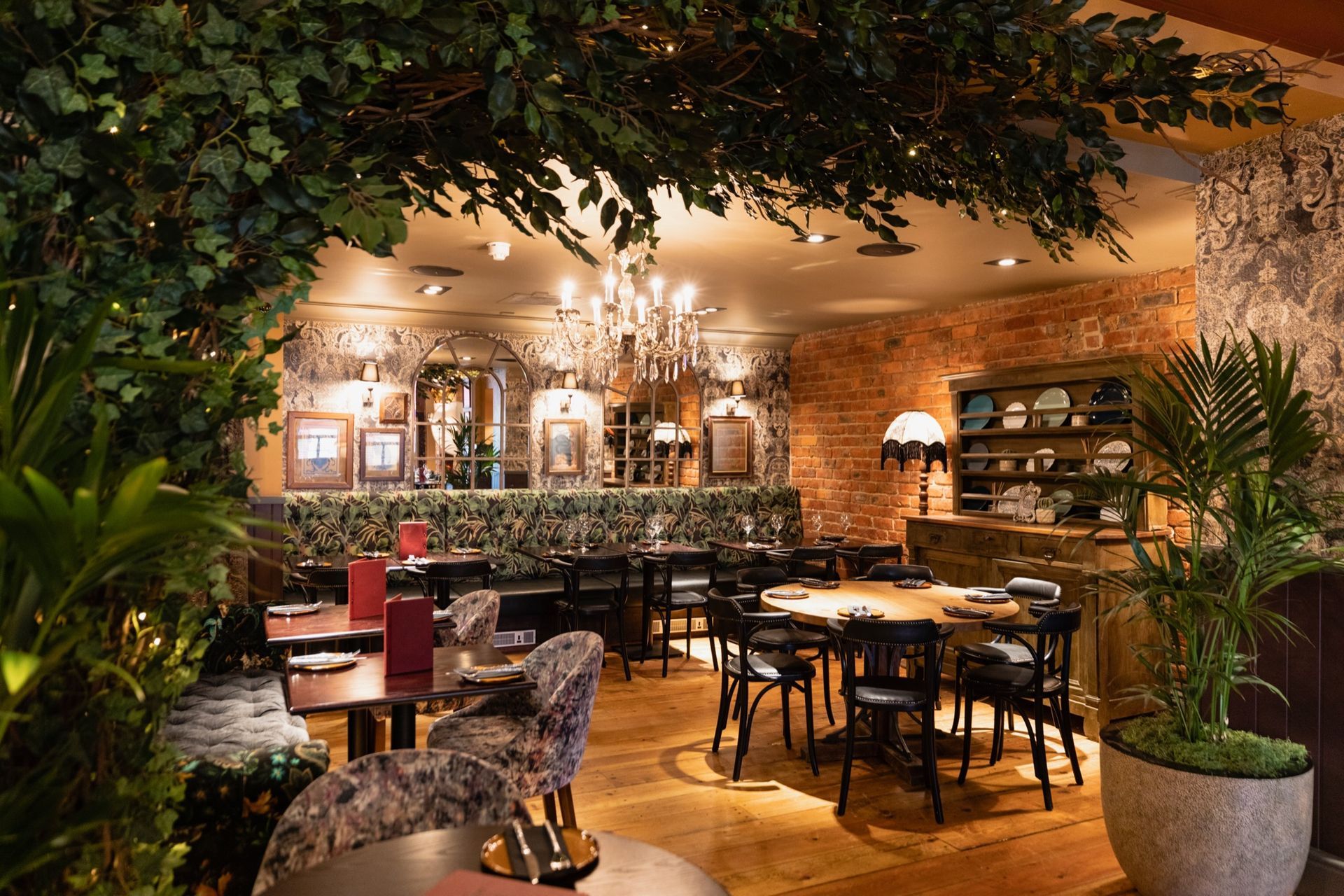 An upscale restaurant interior with rustic brick walls, hanging greenery, a crystal chandelier, and dining furniture.