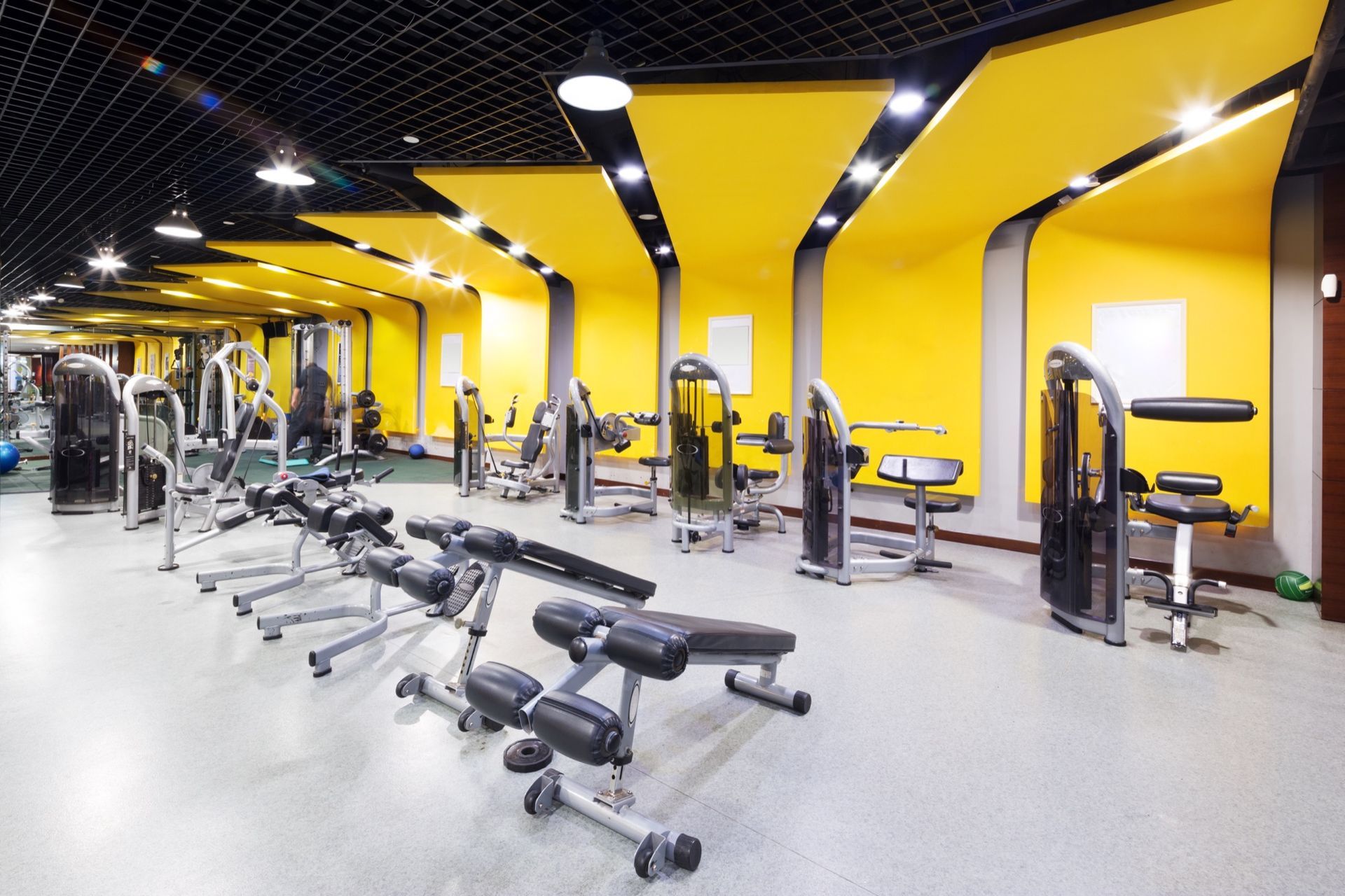 A spacious gym with rows of weight machines and exercise benches, featuring yellow wall panels and a black grid ceiling.