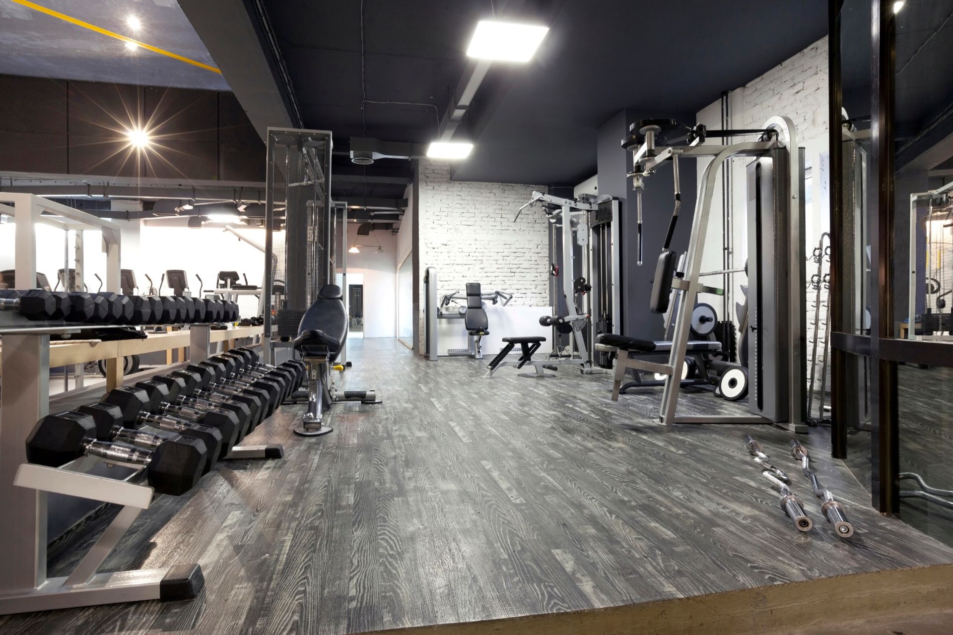 A modern fitness gym with a dumbbell rack in the foreground and weight training equipment on a grey wood-look floor.