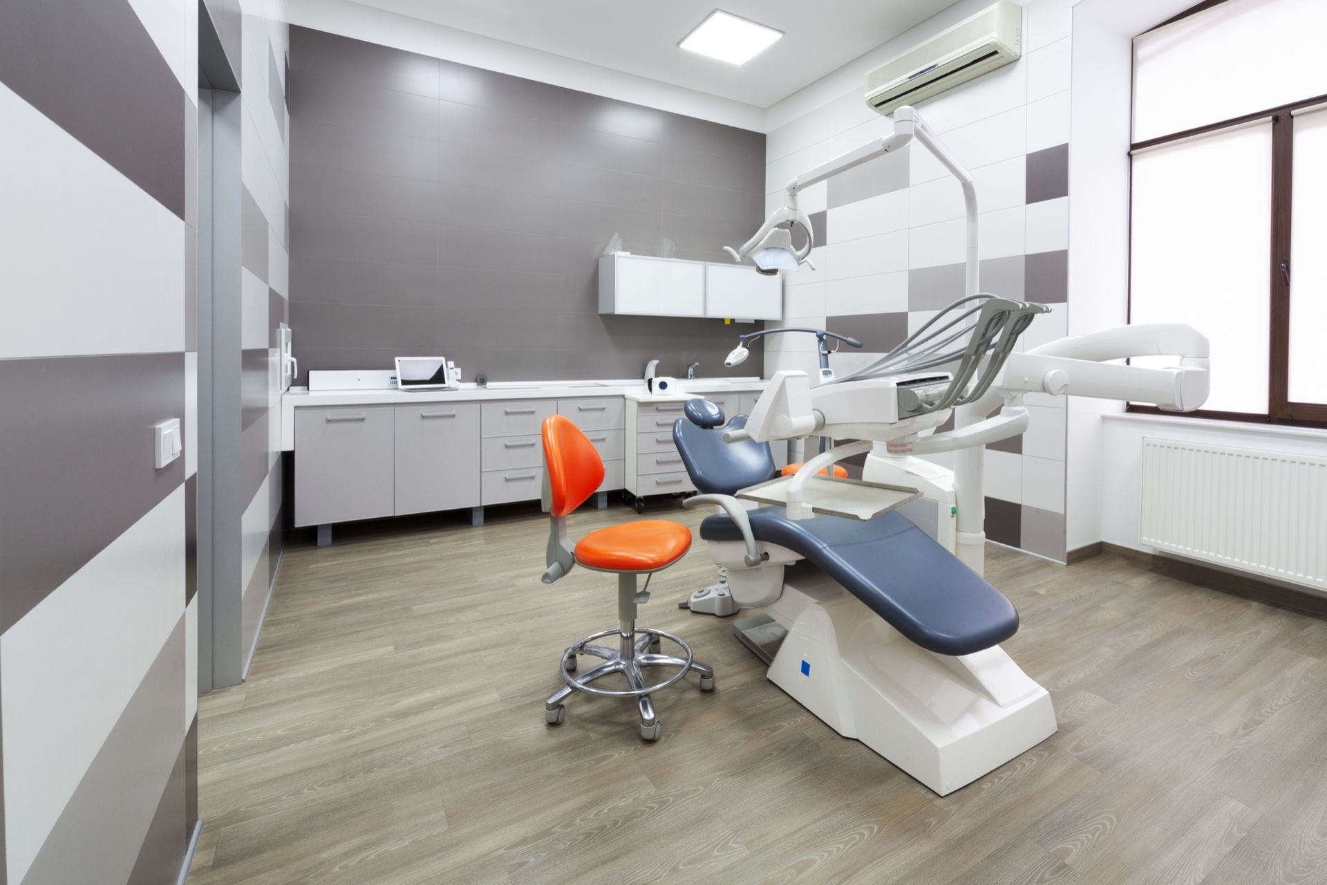 A bright, modern dental office with a blue patient chair, an orange swivel stool, white cabinets, and gray tiled walls.