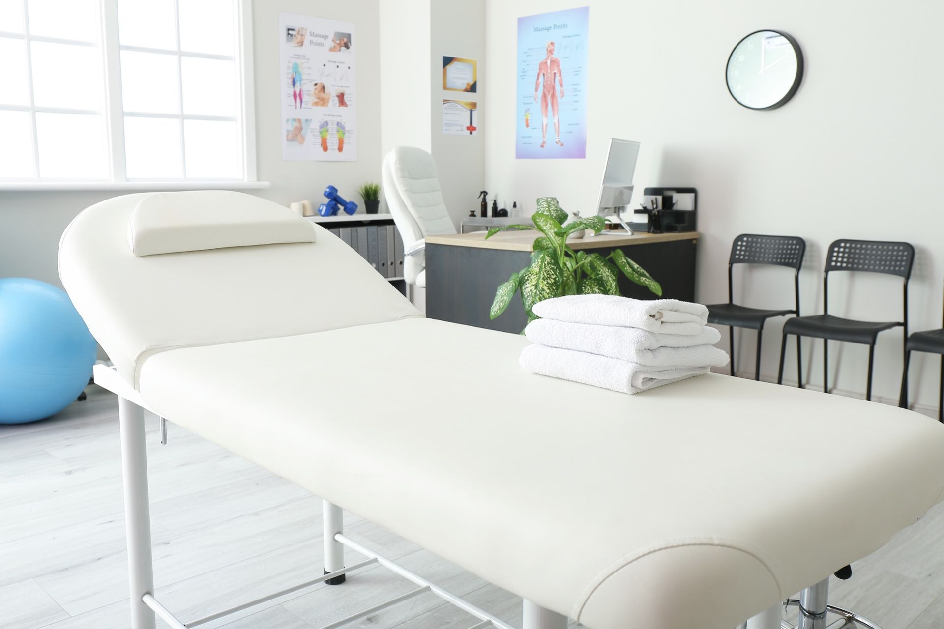 A white treatment table with folded towels sits in a clinic office featuring a desk, chairs, and anatomical posters.