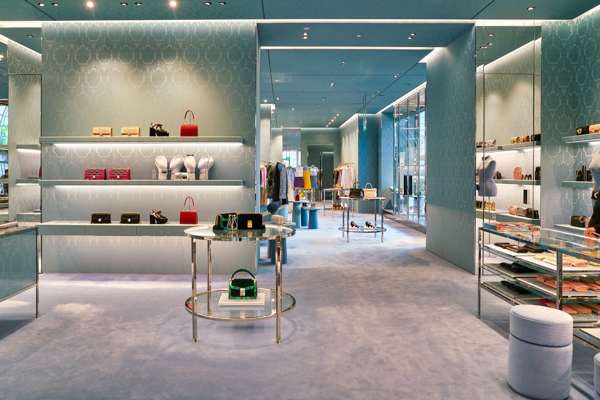 A luxury retail store interior with teal walls, illuminated shelves displaying handbags, and a central display table.