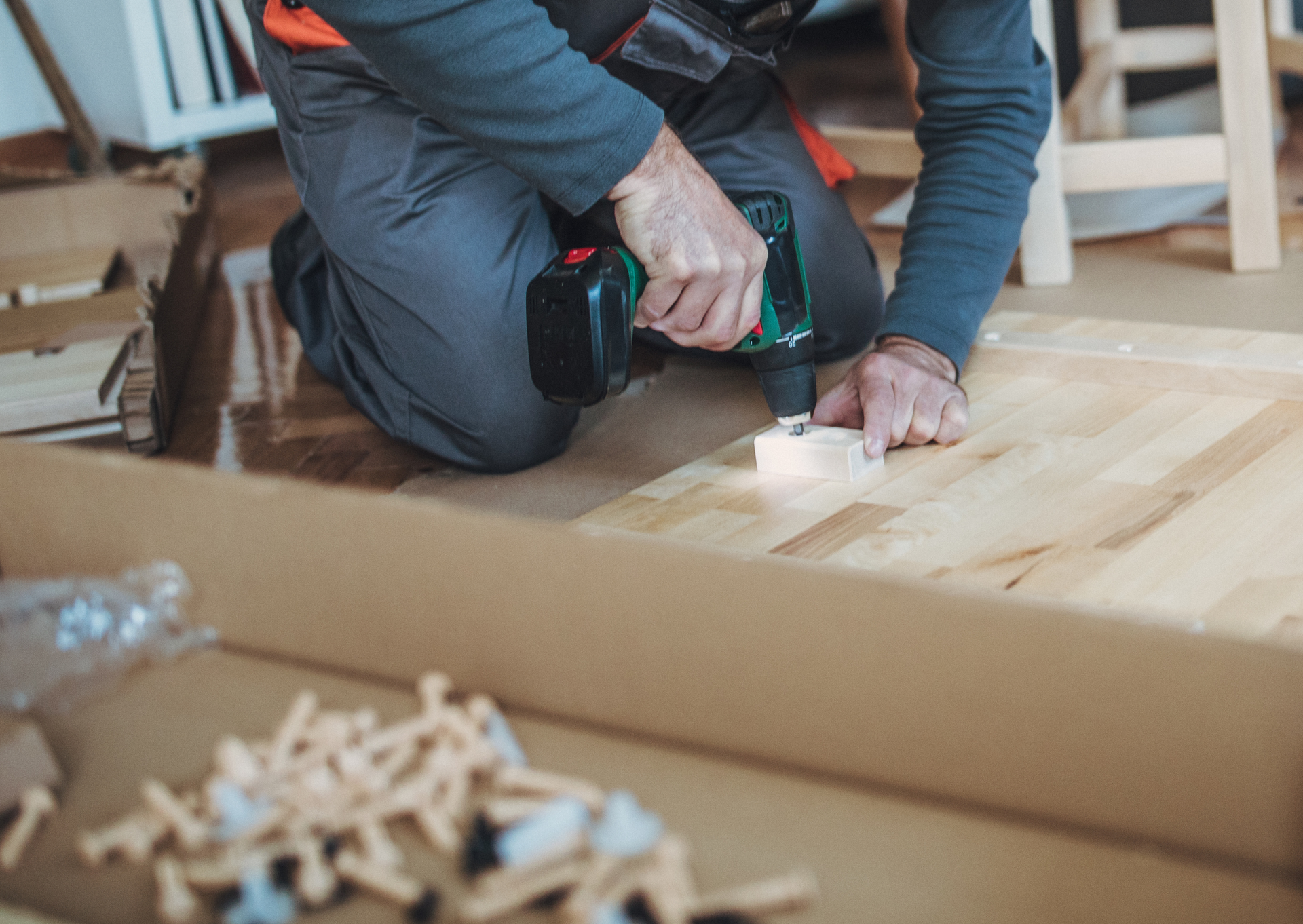 A man is kneeling down while using a drill on a wooden table.