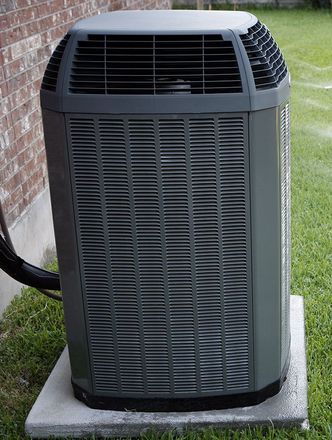 A gray air conditioner is sitting on a concrete platform in front of a brick building.