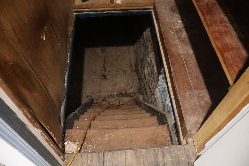 A steep, narrow wooden staircase leading down into a dark, unfinished basement with stone walls.