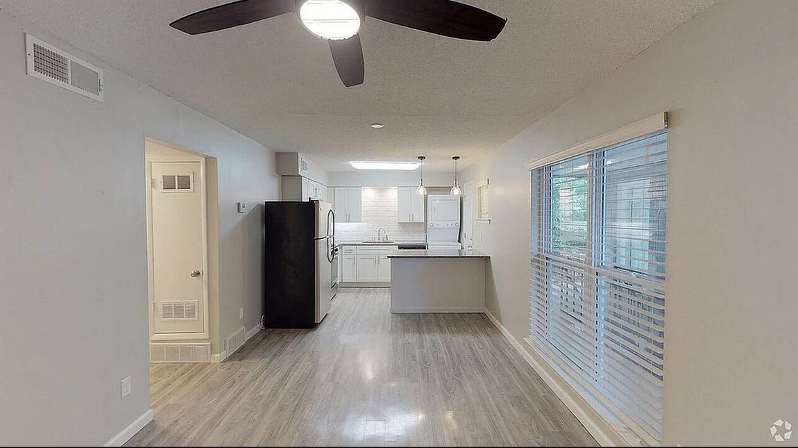 Bright open kitchen and living area with white walls, ceiling fan, and large windows with blinds.