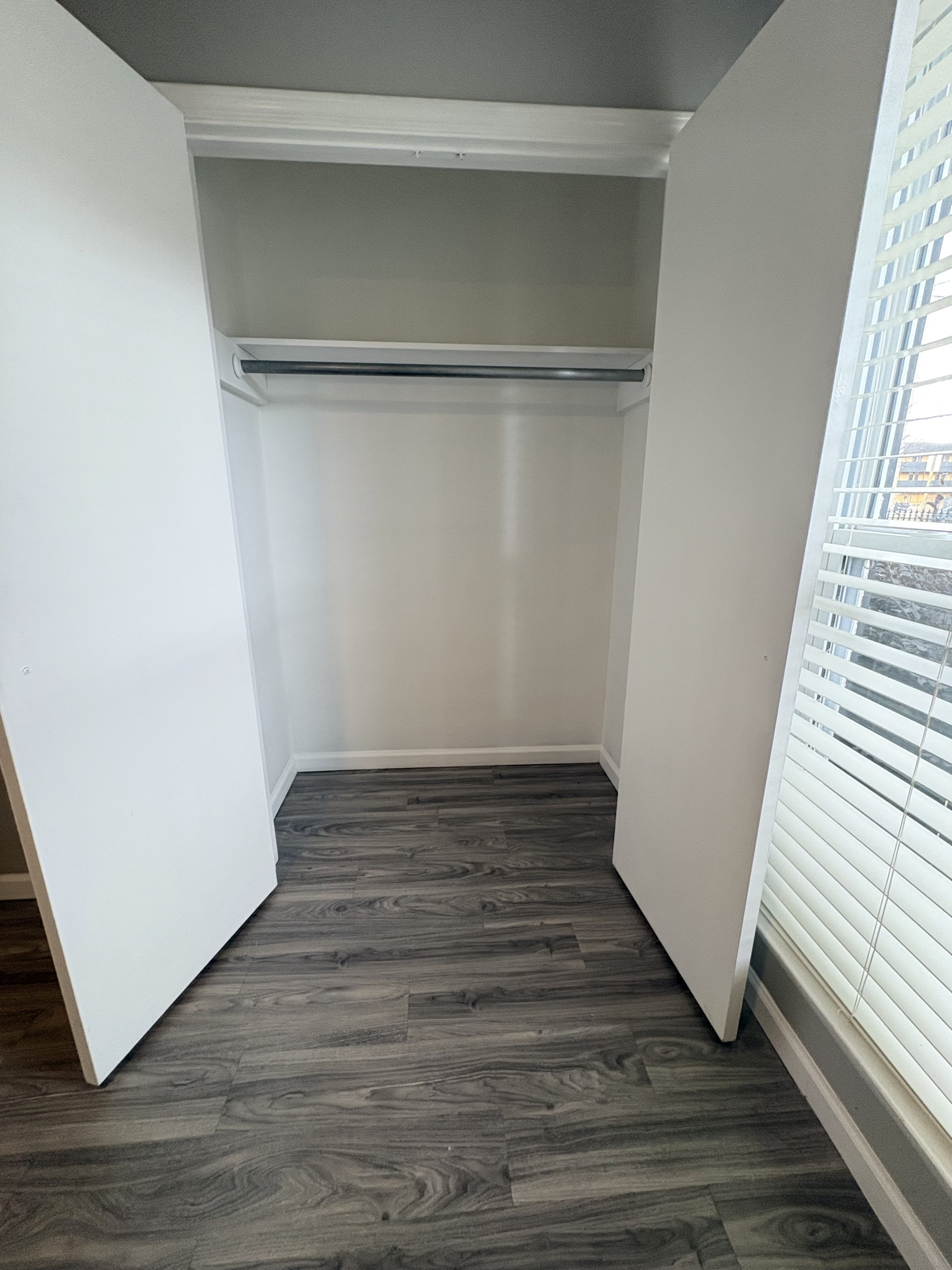 Open closet with white doors, gray walls, and dark wood flooring beside a window with blinds