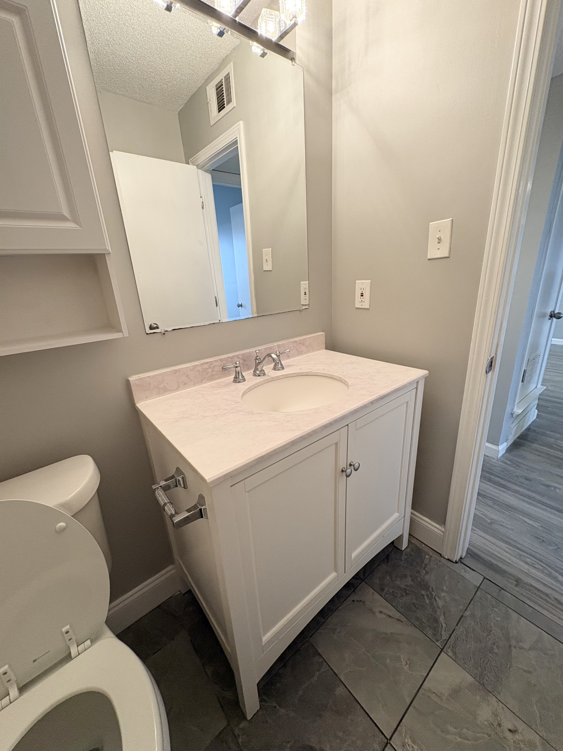 Small bathroom with white vanity, sink, mirror, toilet, and gray tile flooring.
