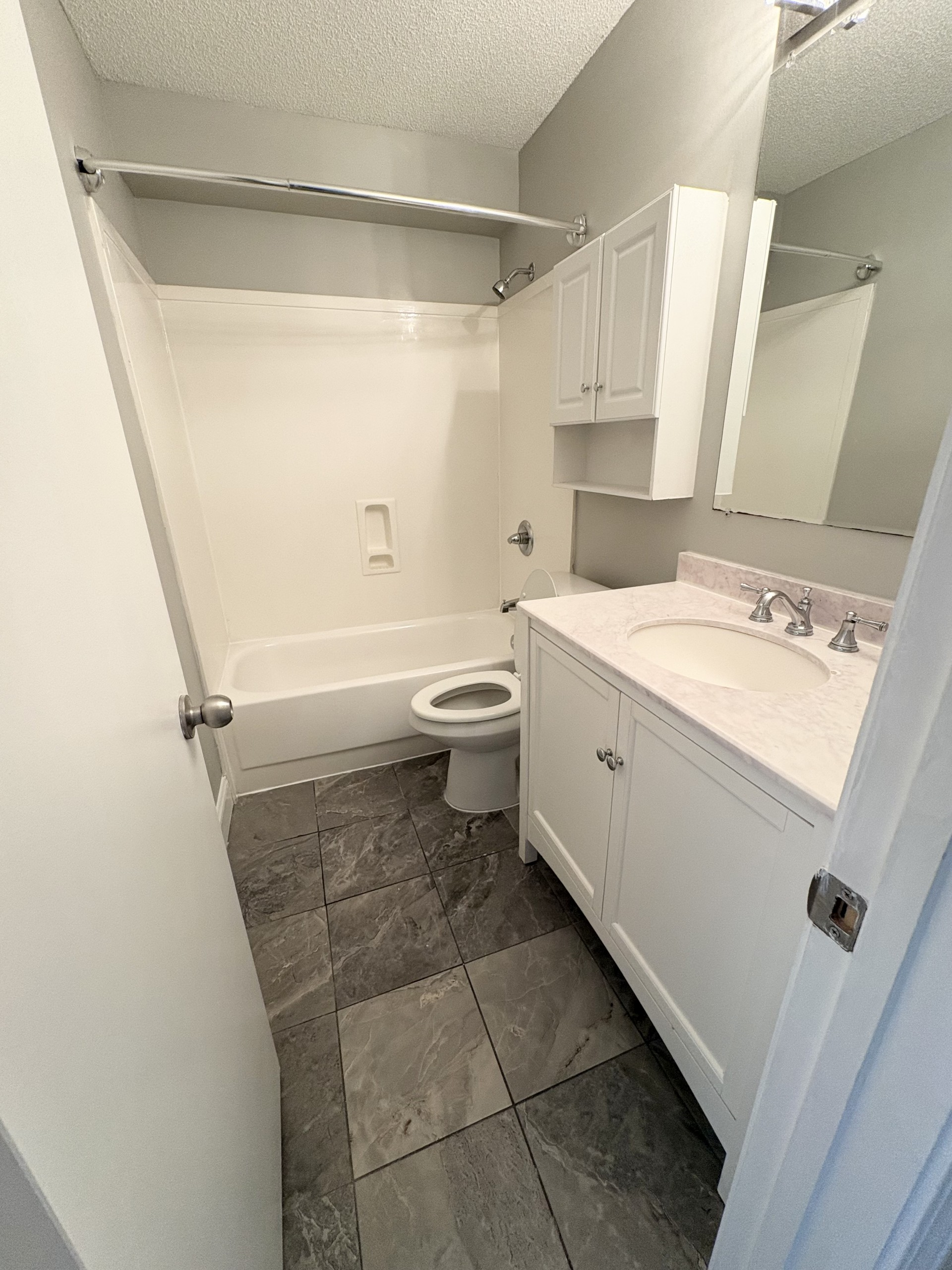 Small bathroom with toilet, vanity sink, mirror, and gray tile floor, viewed from the doorway