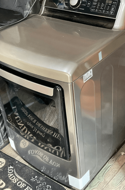 Silver-colored clothes dryer with a front-load door, sitting on a store floor, and partially covered in plastic.