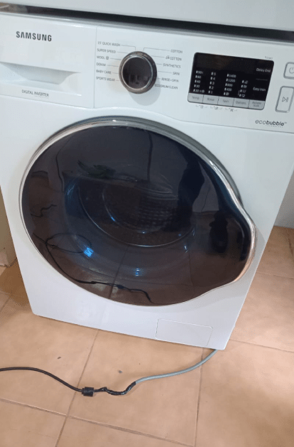 White Samsung washing machine with a black door, in a tiled room.