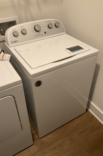 White Whirlpool top-load washing machine next to a dryer. Set in a room with wood-look flooring.