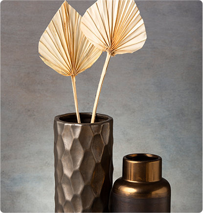 Two dried palm leaf stems in a textured, dark bronze vase, next to a smaller, gold-rimmed vase.
