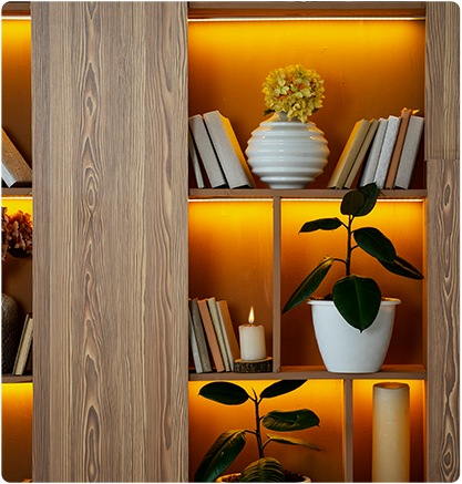 Wooden shelving unit with books, plants, a vase of yellow flowers, and warm lighting.