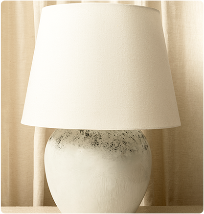 White table lamp with a cream-colored lampshade, set against a cream curtain.