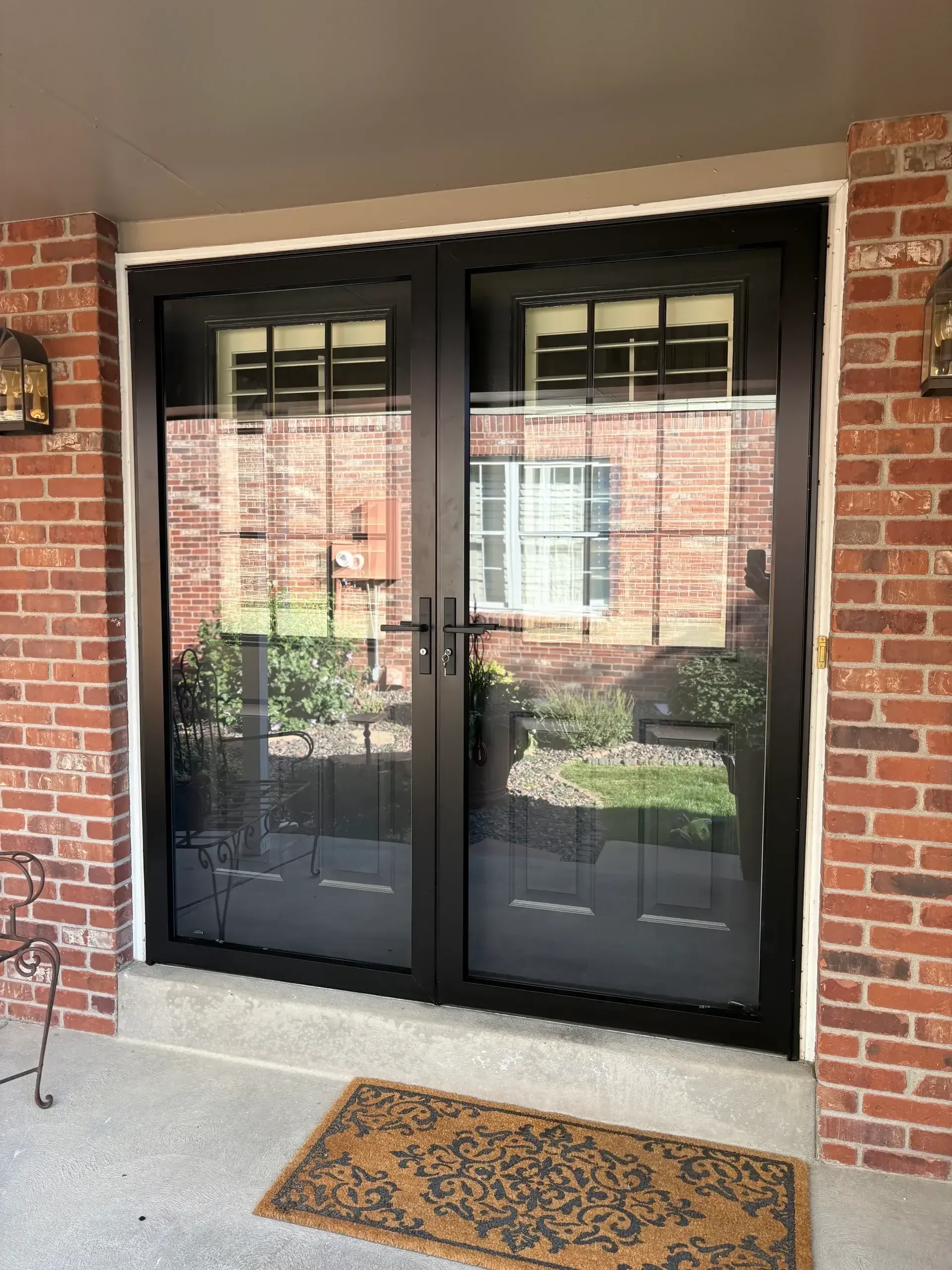 Black double doors with screen, brick walls, a doormat, and light fixtures.