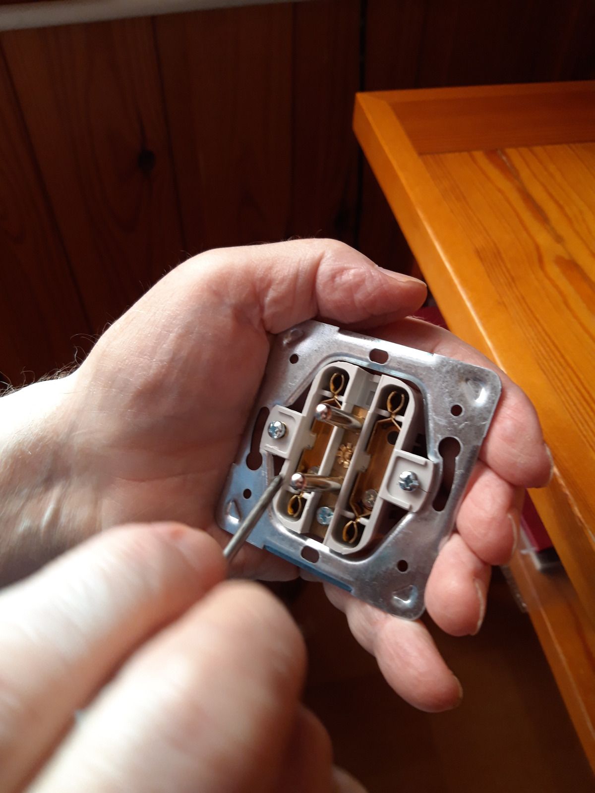 Person holding a light switch, using a screwdriver to make a connection; wooden table in the background.