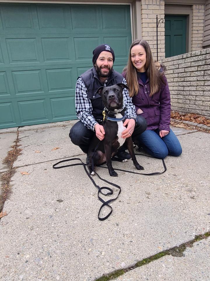 A person and a woman sit on a driveway with a black-and-white dog on a leash in front of a green garage door.