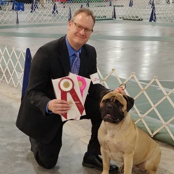 A man in a suit kneeling next to a dog holding a dog training award